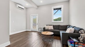 A modern living room with a dark grey sofa, a round wooden coffee table, and a large window with a view of greenery. - Tranquil River Home by Avantstay Blue Ridge Views + Balcony + Two Living Rooms Mins to Downtown (Asheville)