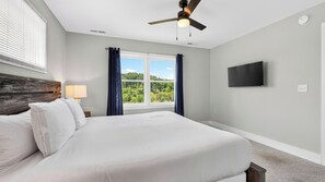 A bedroom with a bed, a ceiling fan, a window with curtains, a wall-mounted TV, and a nightstand with a lamp. - Tranquil River Home by Avantstay Blue Ridge Views + Balcony + Two Living Rooms Mins to Downtown (Asheville)