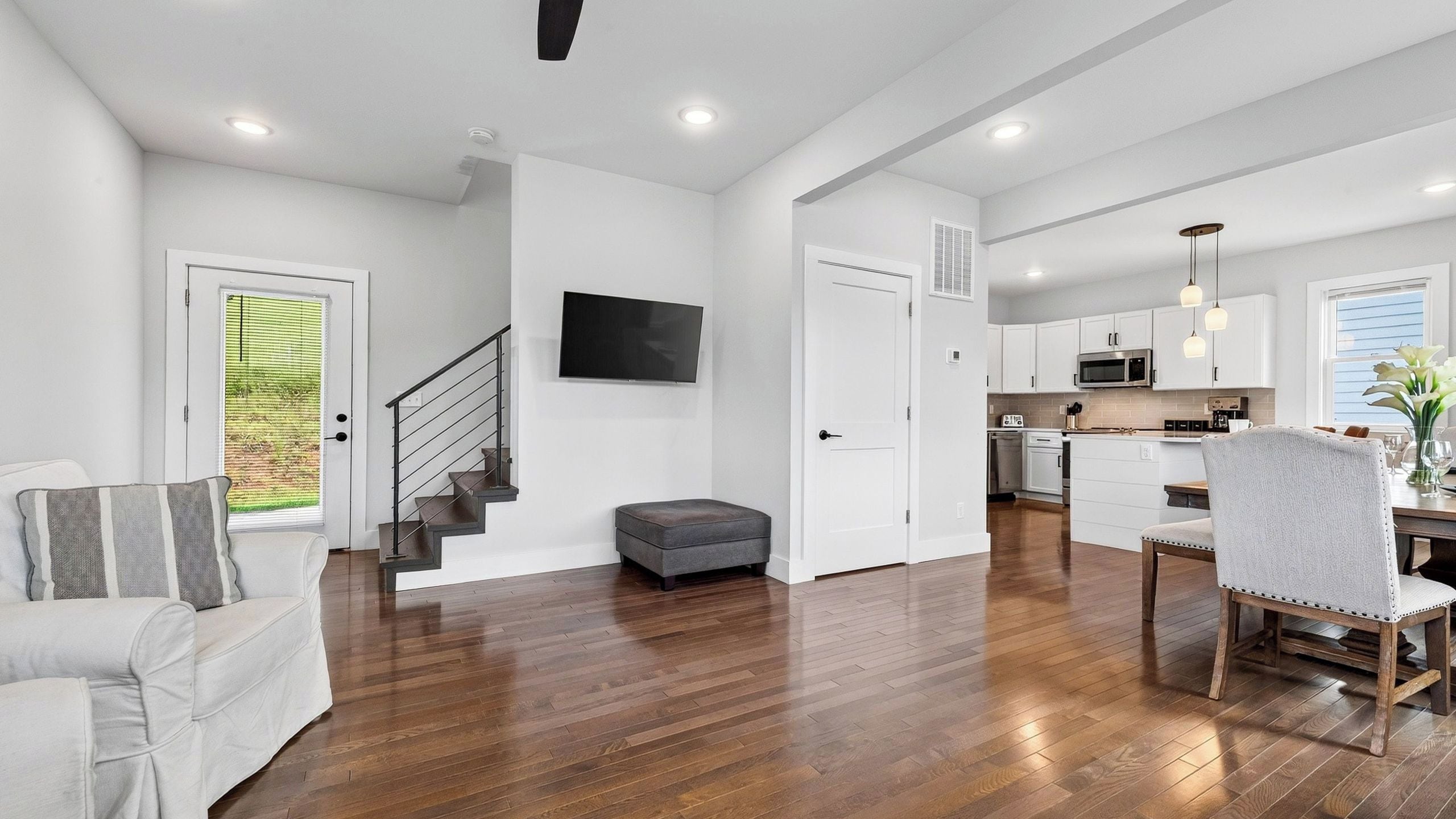 A modern interior with a staircase, a flat-screen TV, a dining area with a table and chairs, and a kitchen with a microwave.