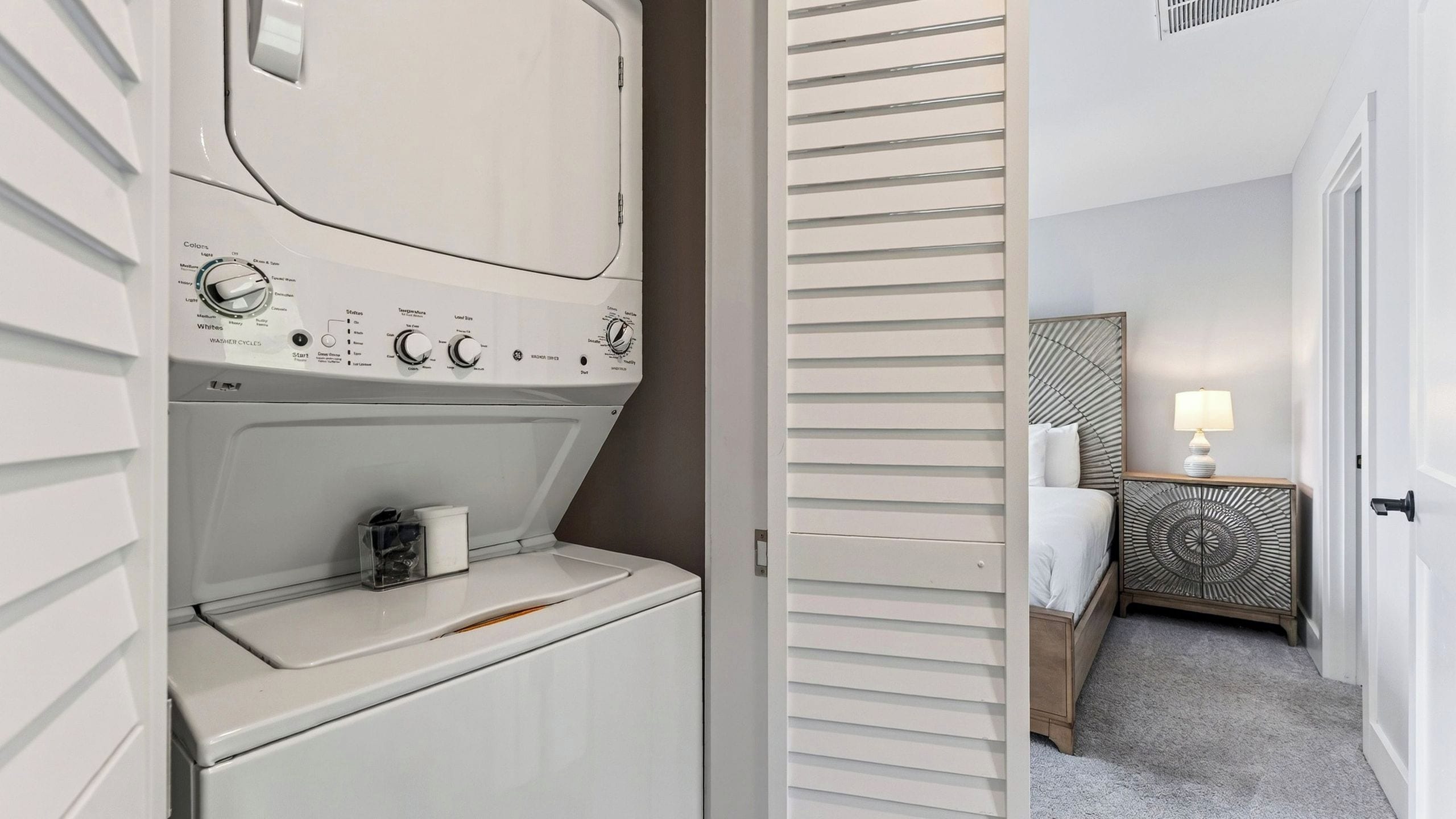 A hotel room with a built-in washer and dryer, a bedside table with a lamp, and a bed with a headboard.