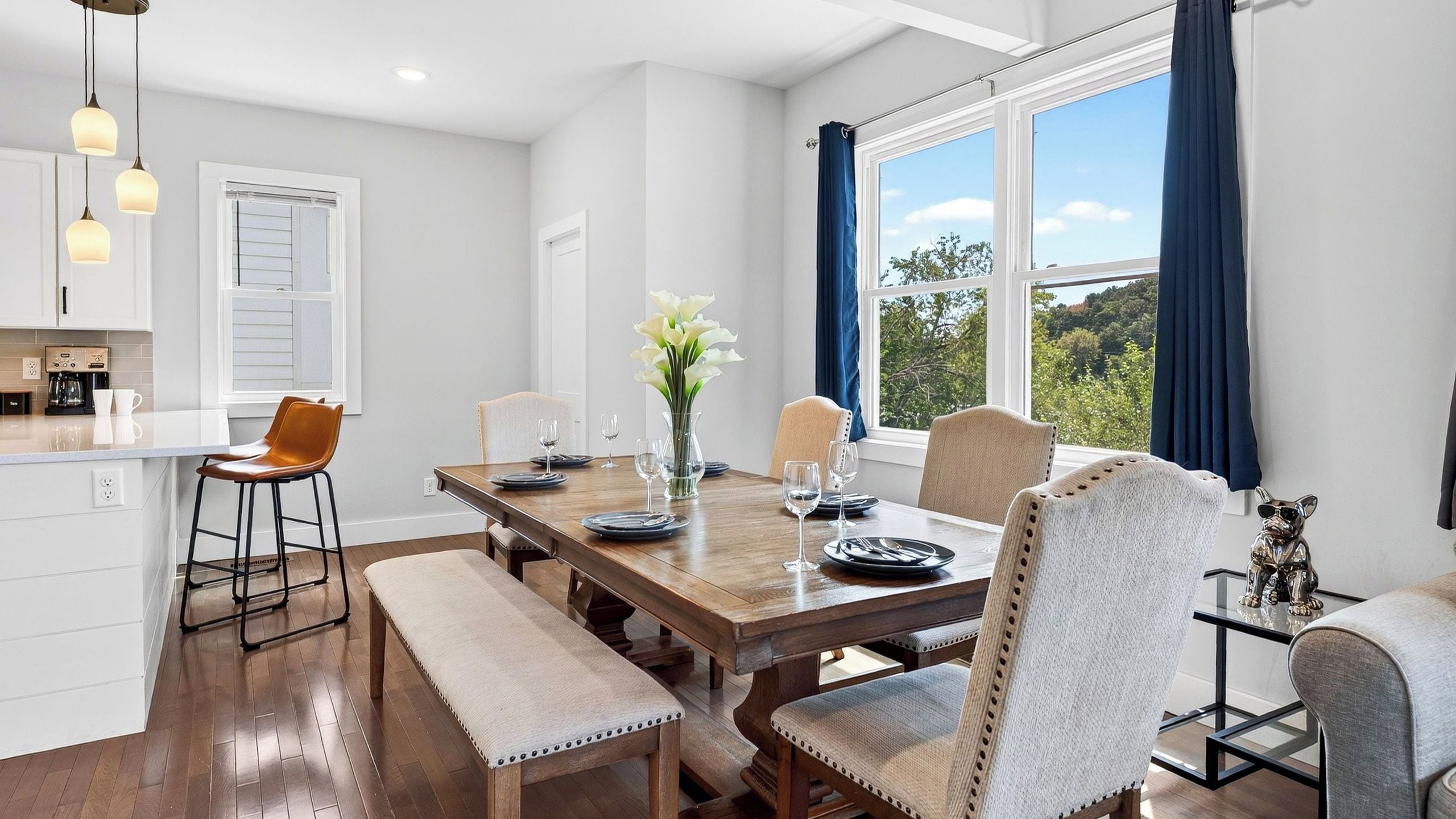 A dining area with a wooden table set for four, a bench, and a view of the outdoors through large windows.