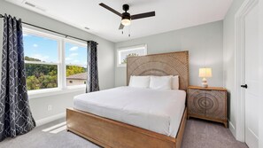 A bedroom with a bed, a nightstand, a wall-mounted headboard, a ceiling fan, and a window with curtains.