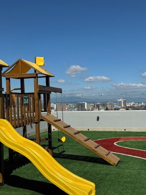 Un parque infantil con un tobogán amarillo y una estructura para escalar, con un paisaje urbano de fondo.