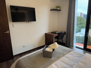 A hotel room with a bed, a desk, a television, and a window with a view of buildings. - Luxury One-Bedroom, City Views, Pool & A/C (Ciudad de Guatemala)