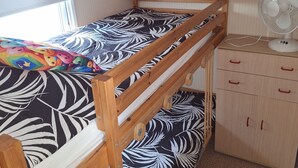 Een stapelbed met een bedspread met een patroon, een houten dressoir met lades, een ventilator en een raam met jaloezieën.