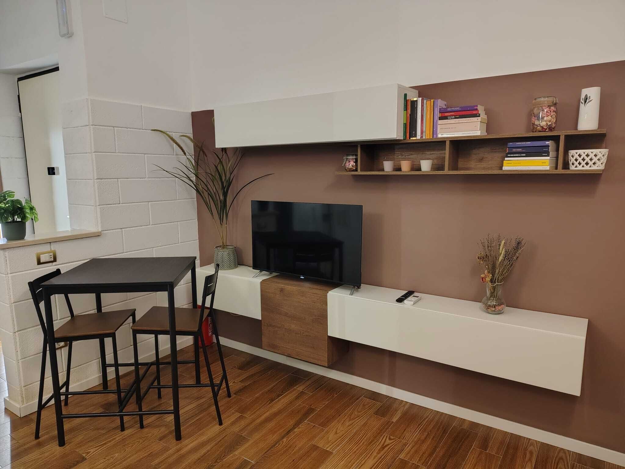 Een moderne woonkamer met een flatscreen televisie, een eettafel met stoelen, een potplant en een boekenkast met boeken en decoratieve objecten.