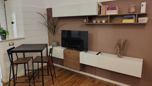 Una sala de estar moderna con un televisor de pantalla plana, una mesa de comedor con sillas, una planta en maceta y una estantería con libros y objetos decorativos.