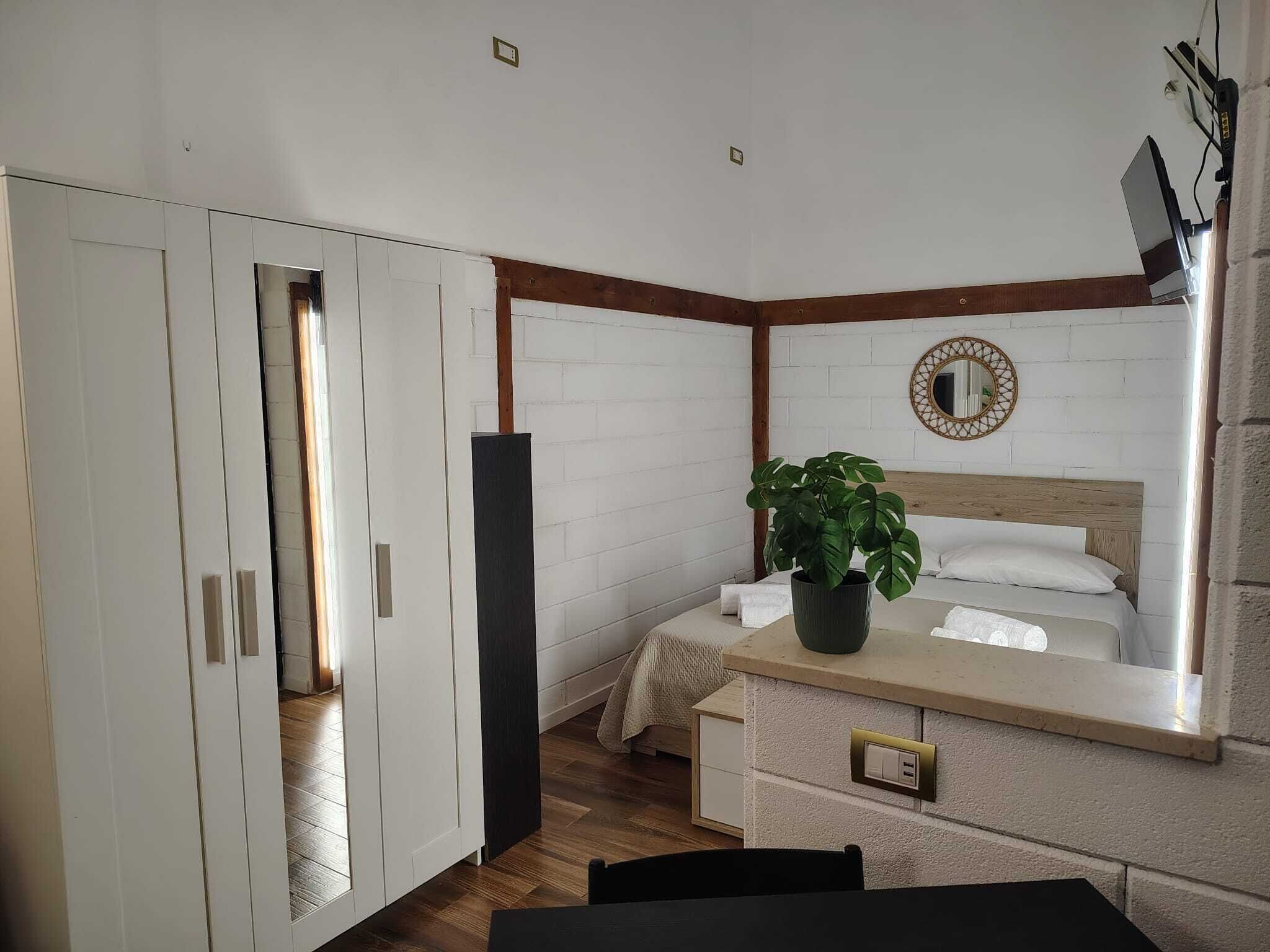 Quarto de hotel com cama, mesa de cabeceira, planta em vaso e espelho na parede.