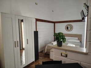 Una habitación de hotel con cama, mesita de noche, planta en maceta y espejo en la pared.