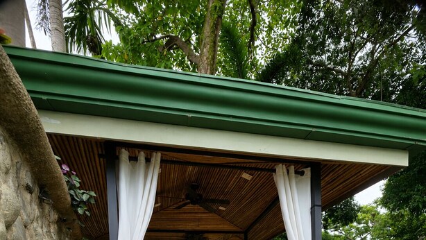 Um deck de madeira com pérgola, cortinas brancas e uma parede de pedra rústica.