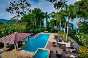 A pool area with lounge chairs and a wooden deck, surrounded by tropical vegetation and overlooking a body of water.
