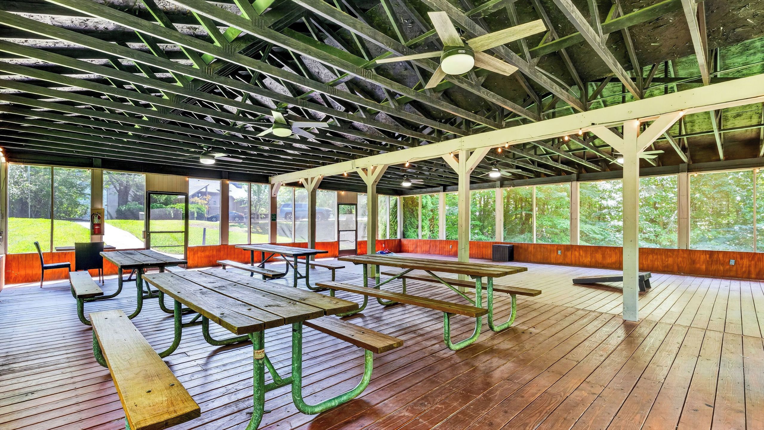 Ein überdachter Picknickbereich im Freien mit Holzbänken und -tischen, einem Deckenventilator und großen Fenstern mit Blick auf die umliegende Natur.