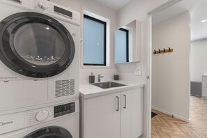 A modern laundry room with a front-loading washing machine, a sink, and a mirror above the counter. - The Cozy Koha - Taupo Holiday Home (Taupo)