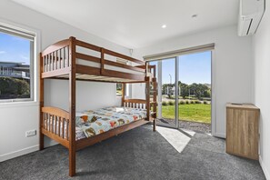 A bunk bed with a lower bunk, a small cabinet, and a large sliding glass door to a garden. - Starling - Mangawhai Heads Holiday Home (Mangawhai)