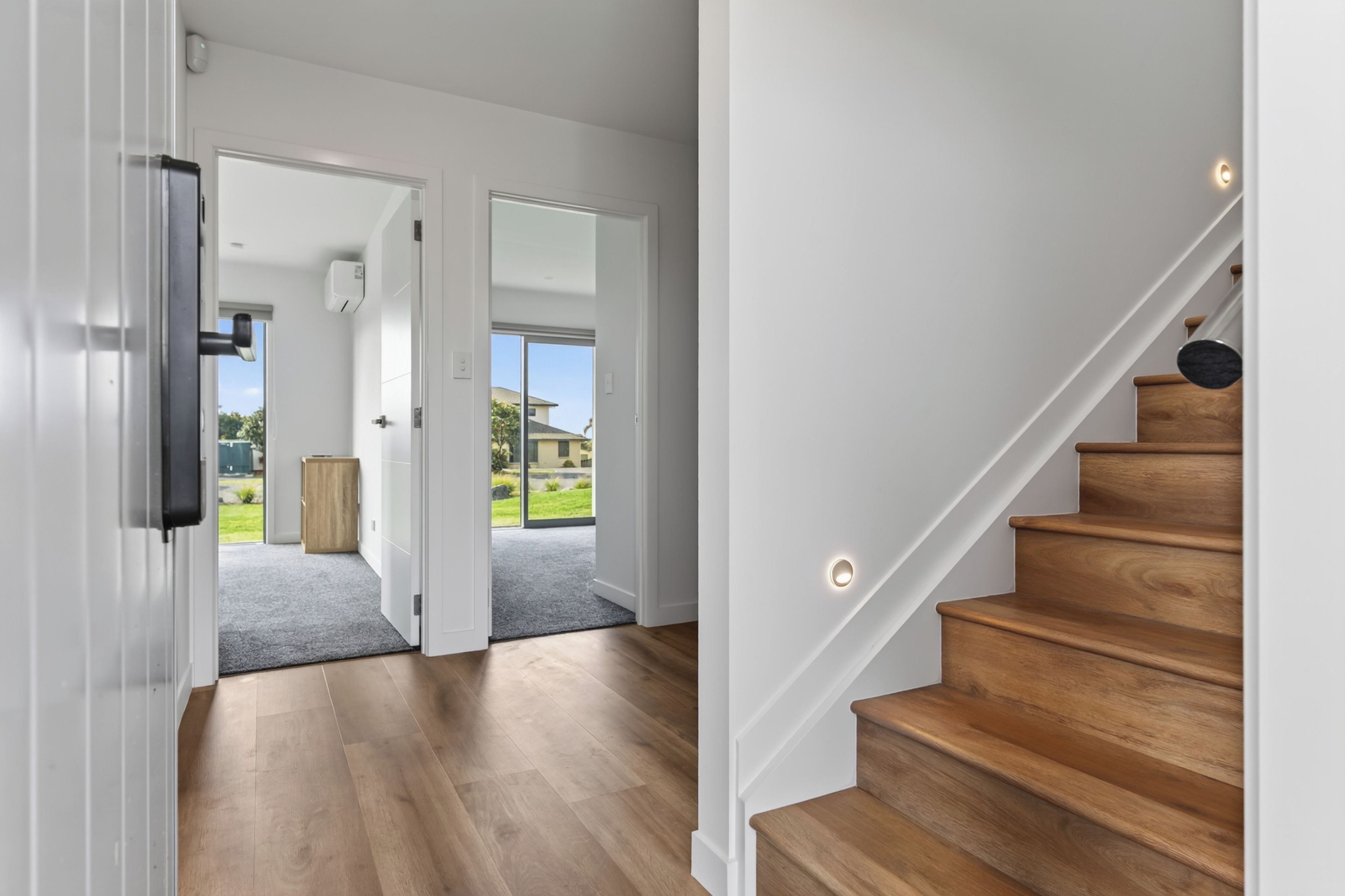 A modern interior with a wooden staircase, a hallway leading to a door, and a view of the outdoors.