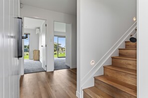 A modern interior with a wooden staircase, a hallway leading to a door, and a view of the outdoors. - Starling - Mangawhai Heads Holiday Home (Mangawhai)