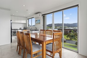 A modern kitchen with white cabinets, a dining table with six wooden chairs, and a large window offering a view of a green landscape and distant hills. - Starling - Mangawhai Heads Holiday Home (Mangawhai)