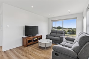 A modern living room with a flat-screen TV, grey sofa, and a large window offering a view of the outdoors. - Starling - Mangawhai Heads Holiday Home (Mangawhai)