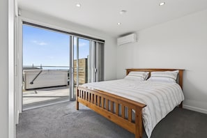 A bedroom with a wooden bed, grey carpet, and a sliding glass door leading to a balcony. - Starling - Mangawhai Heads Holiday Home (Mangawhai)
