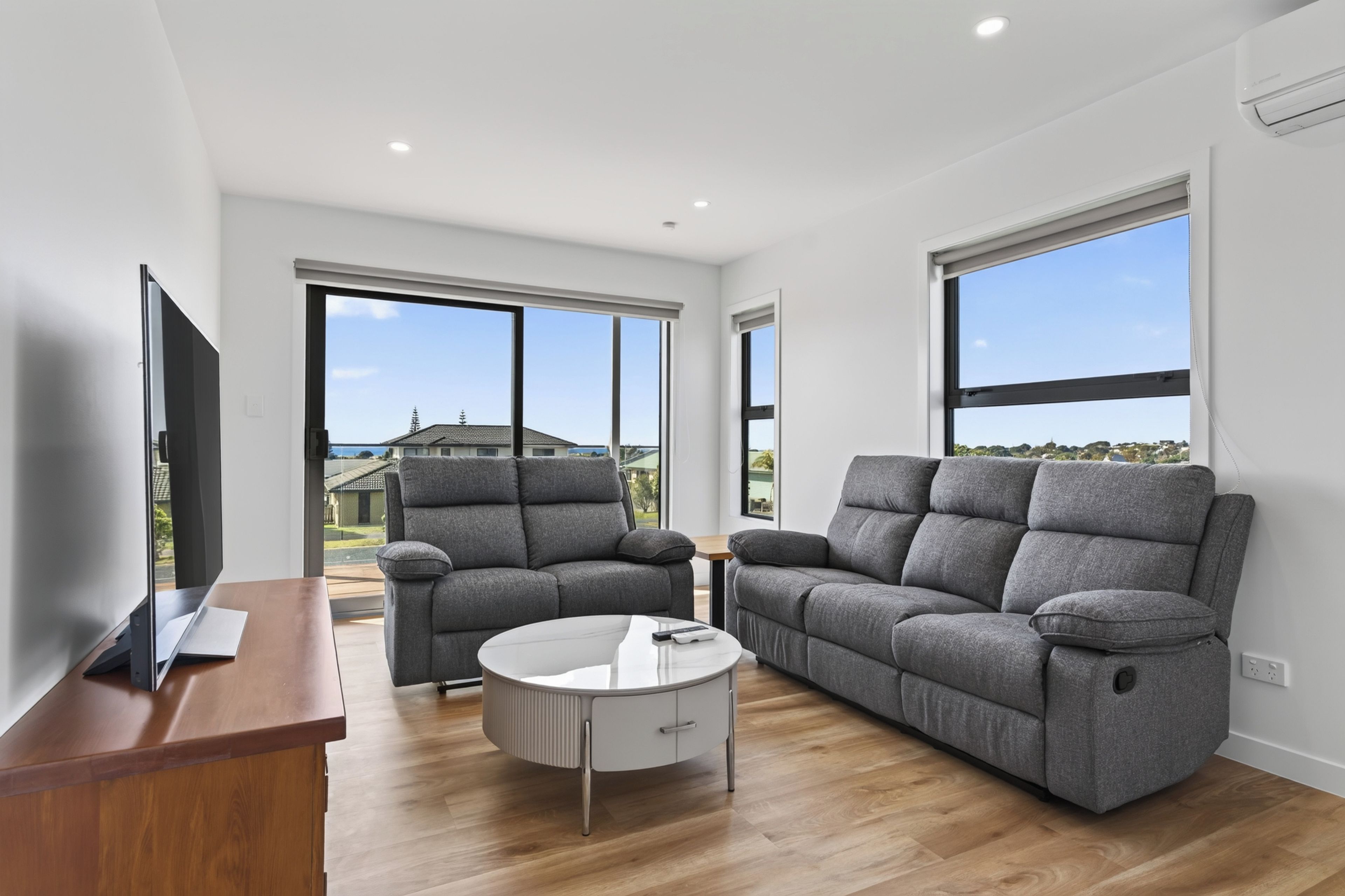 A modern living room with a flat-screen TV, grey sofas, a round coffee table, and large windows offering a view of the outside.