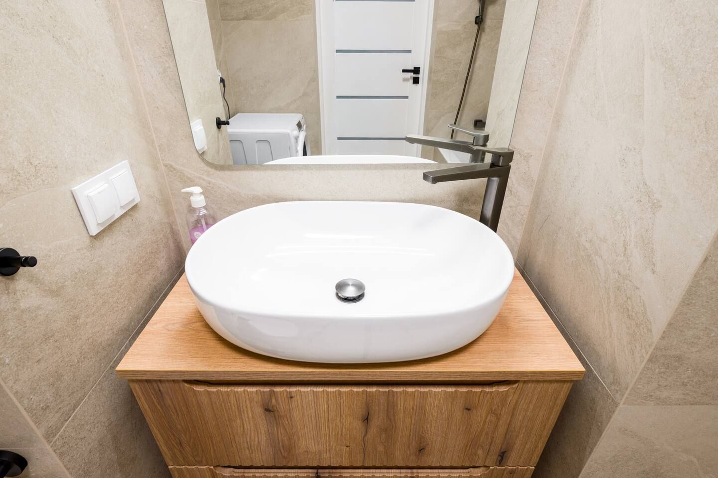 A bathroom with a round white sink on a wooden vanity, a mirror above the sink, and a door in the background.