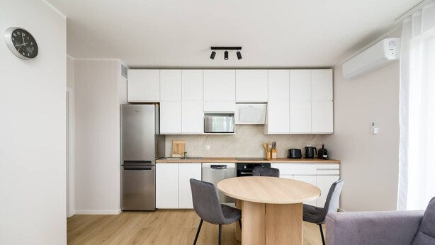 A modern kitchen with white cabinets, a stainless steel refrigerator, a microwave, and a dishwasher. There is a round wooden table with two chairs and a grey sofa in the living area. - Wrocław Three-Bedroom Apt — Parking, A/C & Balcony (Wrocław)