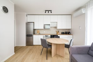 A modern kitchen with white cabinets, a stainless steel refrigerator, a microwave, and a dishwasher. There is a round wooden table with two chairs and a grey sofa in the living area.