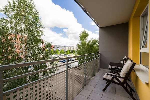 A balcony with a table and chairs, a view of a parking lot, and buildings in the background.