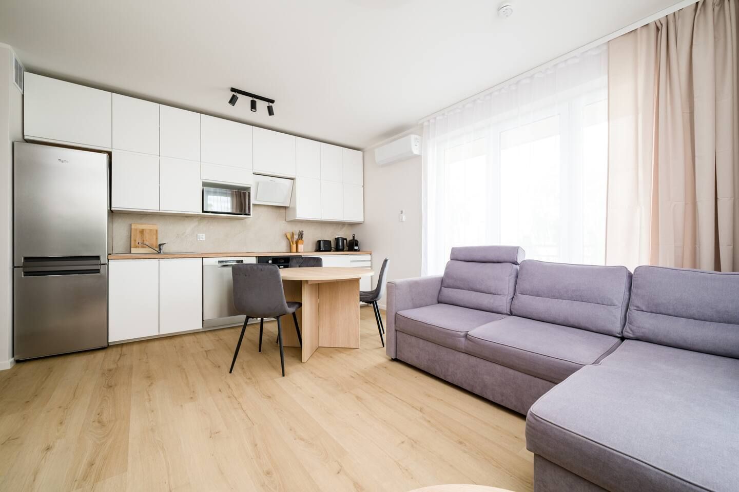 Een moderne keuken met witte kasten, een roestvrijstalen koelkast en een magnetron. Er is een eethoek met een houten tafel en een grijze stoel. Een grijze bank staat in de woonkamer, en de kamer heeft grote ramen met gordijnen.