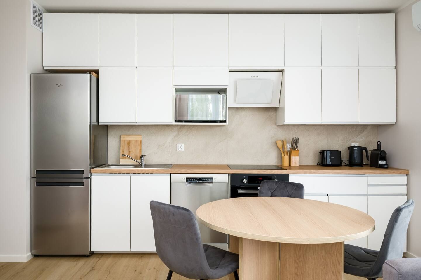 Una cocina moderna con armarios blancos, un refrigerador de acero inoxidable, un microondas y un lavavajillas. Hay una encimera de madera con un fregadero y una mesa redonda con sillas.