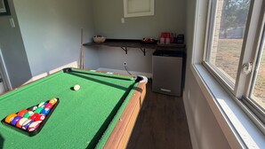 Uma mesa de bilhar com bolas, uma pequena geladeira e uma janela com vista para árvores.