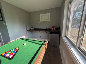 Une table de billard avec des billes, un petit réfrigérateur et une fenêtre donnant sur des arbres.