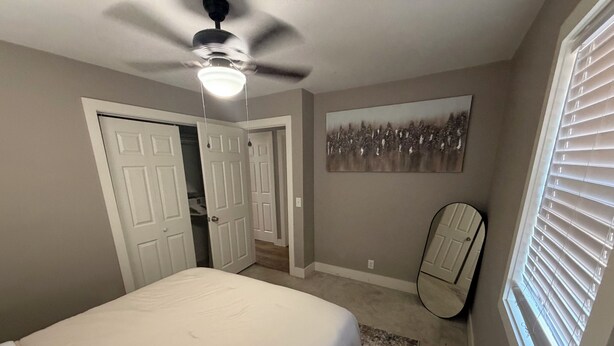 Quarto com cama, ventilador de teto, porta, espelho e janela com persianas.