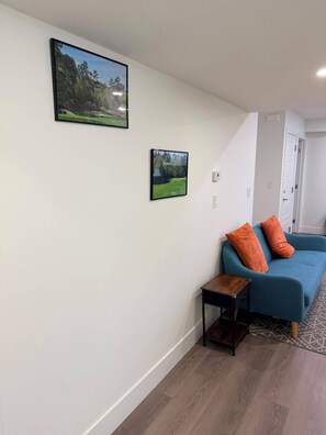 Ein Zimmer mit einem blauen Sofa, zwei orangenen Kissen, einem kleinen Holztisch und zwei gerahmten Bildern an der Wand.