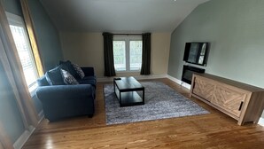 Ein geräumiges Wohnzimmer mit Holzboden, einem blauen Sofa, einem Couchtisch, einem Fernseher auf einer Holzeinrichtung und einem Fenster mit Vorhängen.