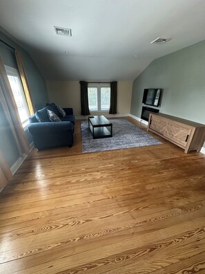 A spacious living room with hardwood flooring, a blue sofa, a coffee table, a TV on a wooden cabinet, and a window with curtains. -  3 BR apartment with central AC in the village. Steps to Main St. (Westhampton Beach)