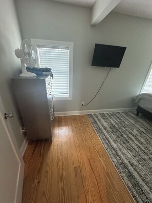 Une pièce avec un plancher en bois, un téléviseur fixé au mur, un ventilateur sur pied et une fenêtre avec des stores.