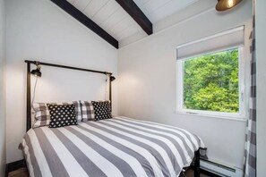 A bed with a striped bedspread and multiple pillows, a window with a view of greenery, and a ceiling with exposed beams. - Stylish 2 Bedroom in Trees. On Free Bus Route (Breckenridge)