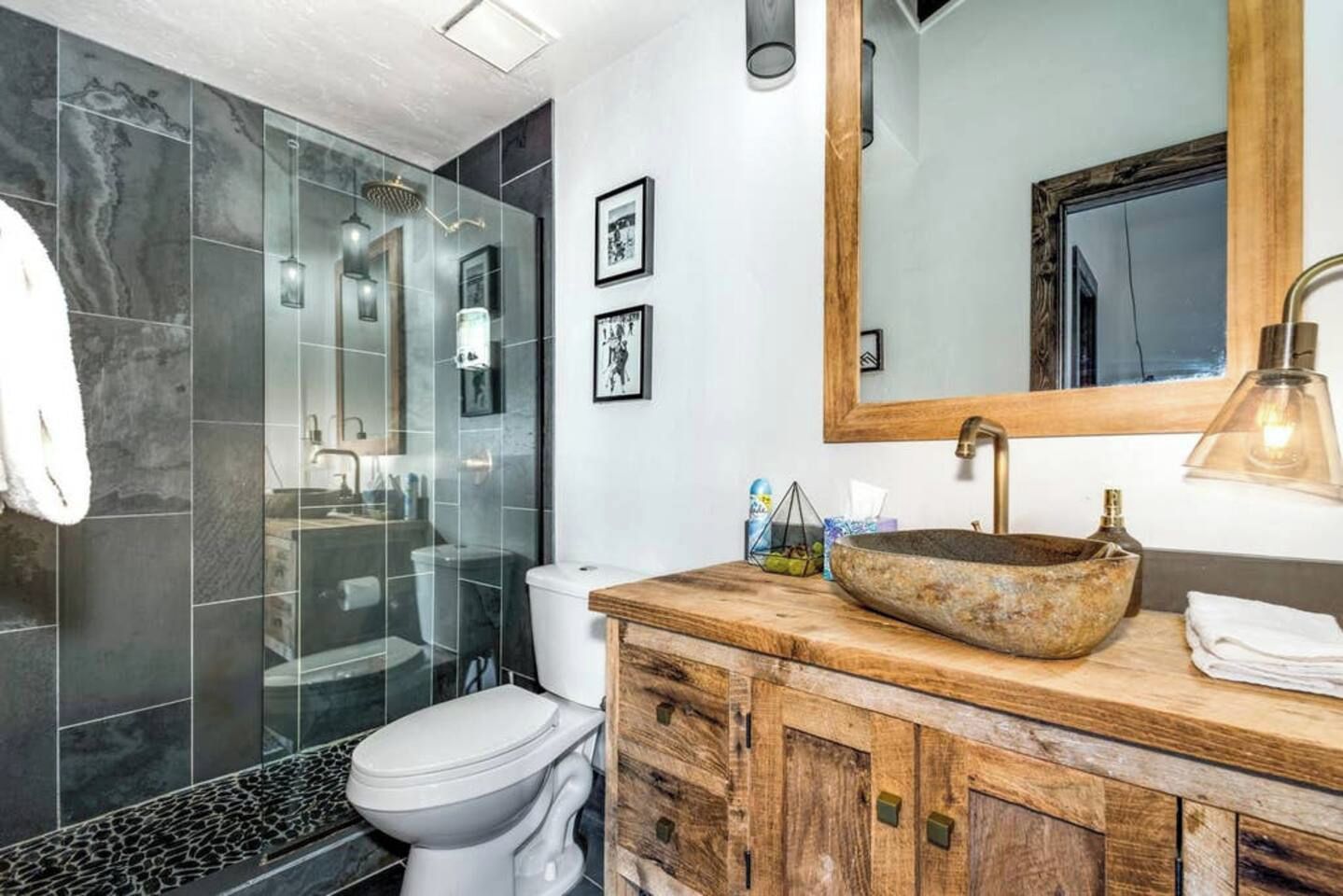 A bathroom with a glass shower enclosure, a white toilet, a wooden vanity with a large vessel sink, and a mirror above the sink.