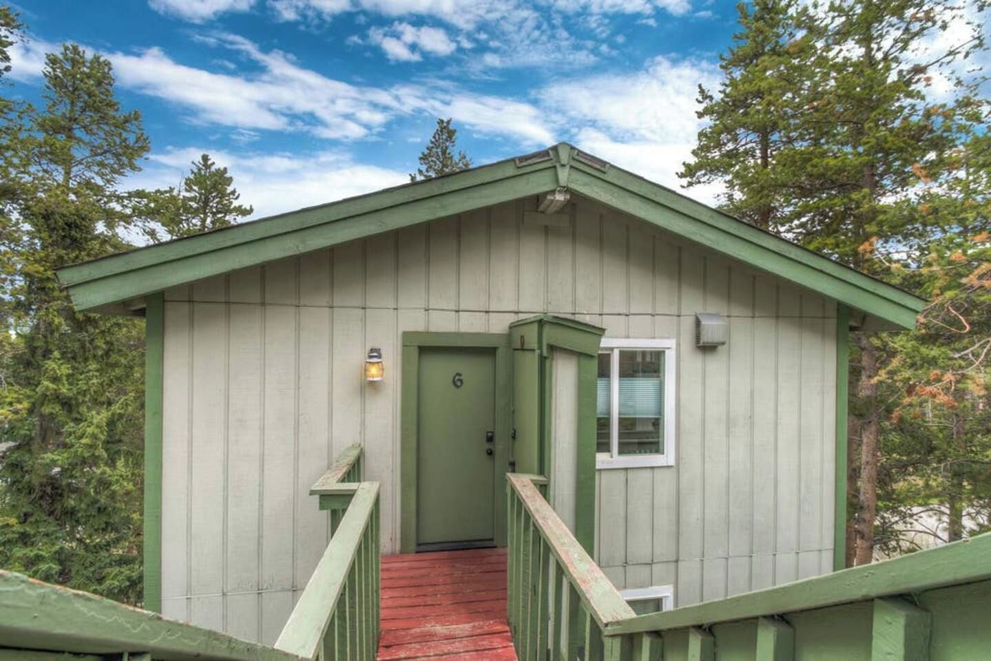 A small, single-story cabin with a green exterior, a red wooden staircase, and a door marked with the number 6.