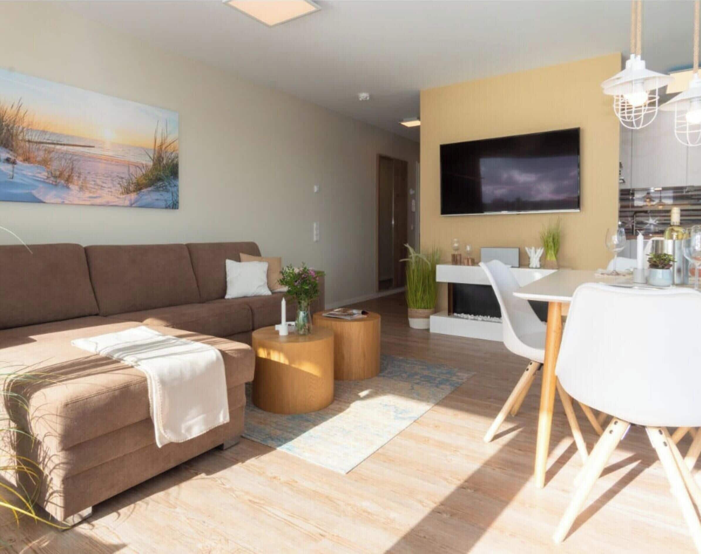 Un salon moderne avec un canapé marron, un téléviseur à écran plat, un coin repas avec une table et des chaises blanches, et une cuisine en arrière-plan.