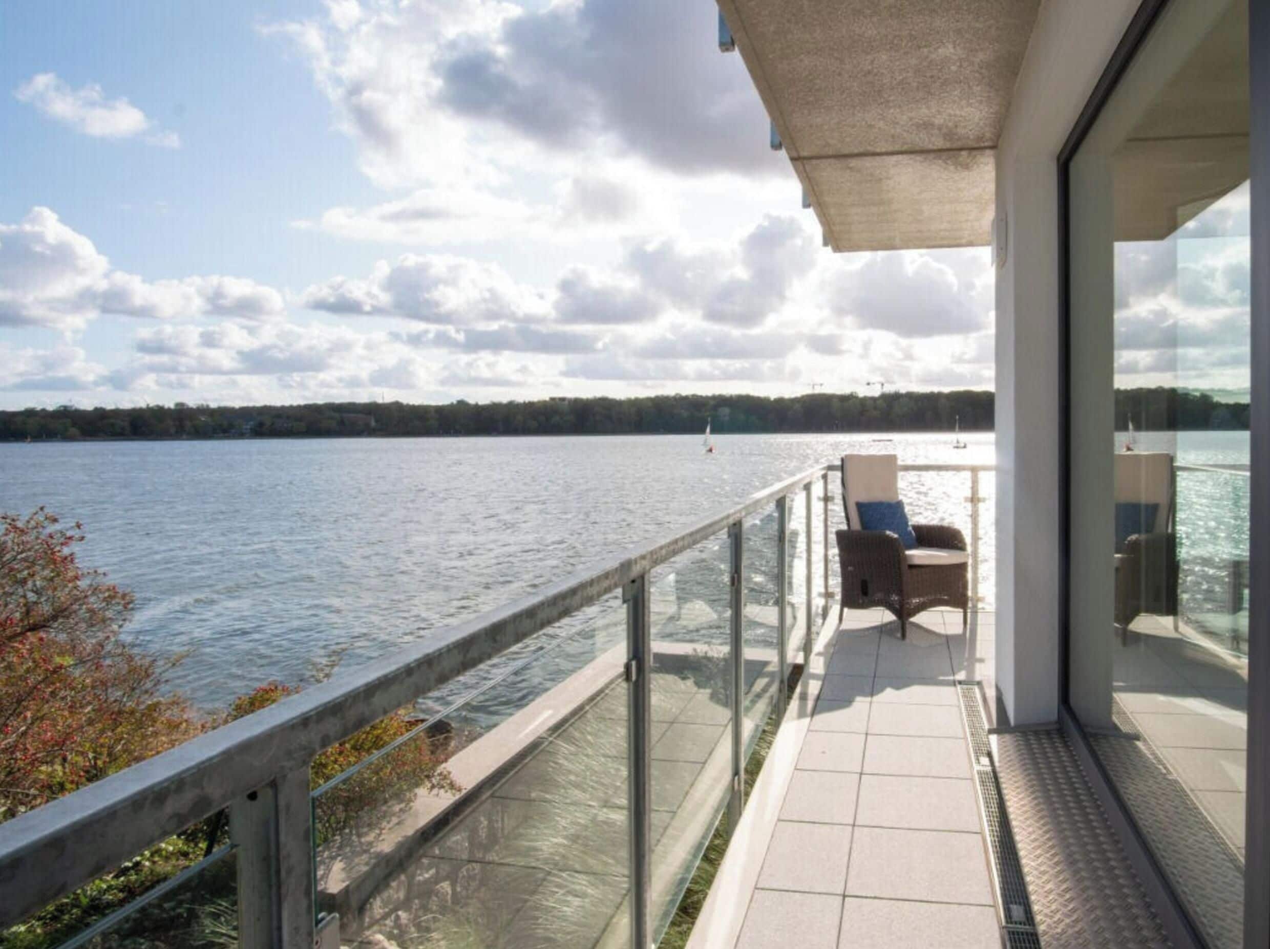 A balcony with a glass railing overlooking a lake, a chair, and a view of the water.
