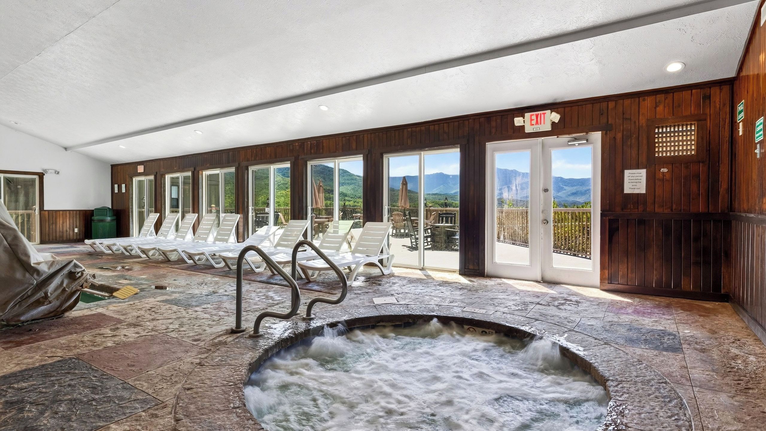 Ein Wellnessbereich mit Whirlpool, Liegestühlen und Blick auf Berge durch große Fenster.