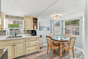 A kitchen with a dining area, wooden cabinets, a coffee maker, and a view of the outdoors. - Saltwater Shanty GolfCart•Linens•OceanLakes (Horry County)
