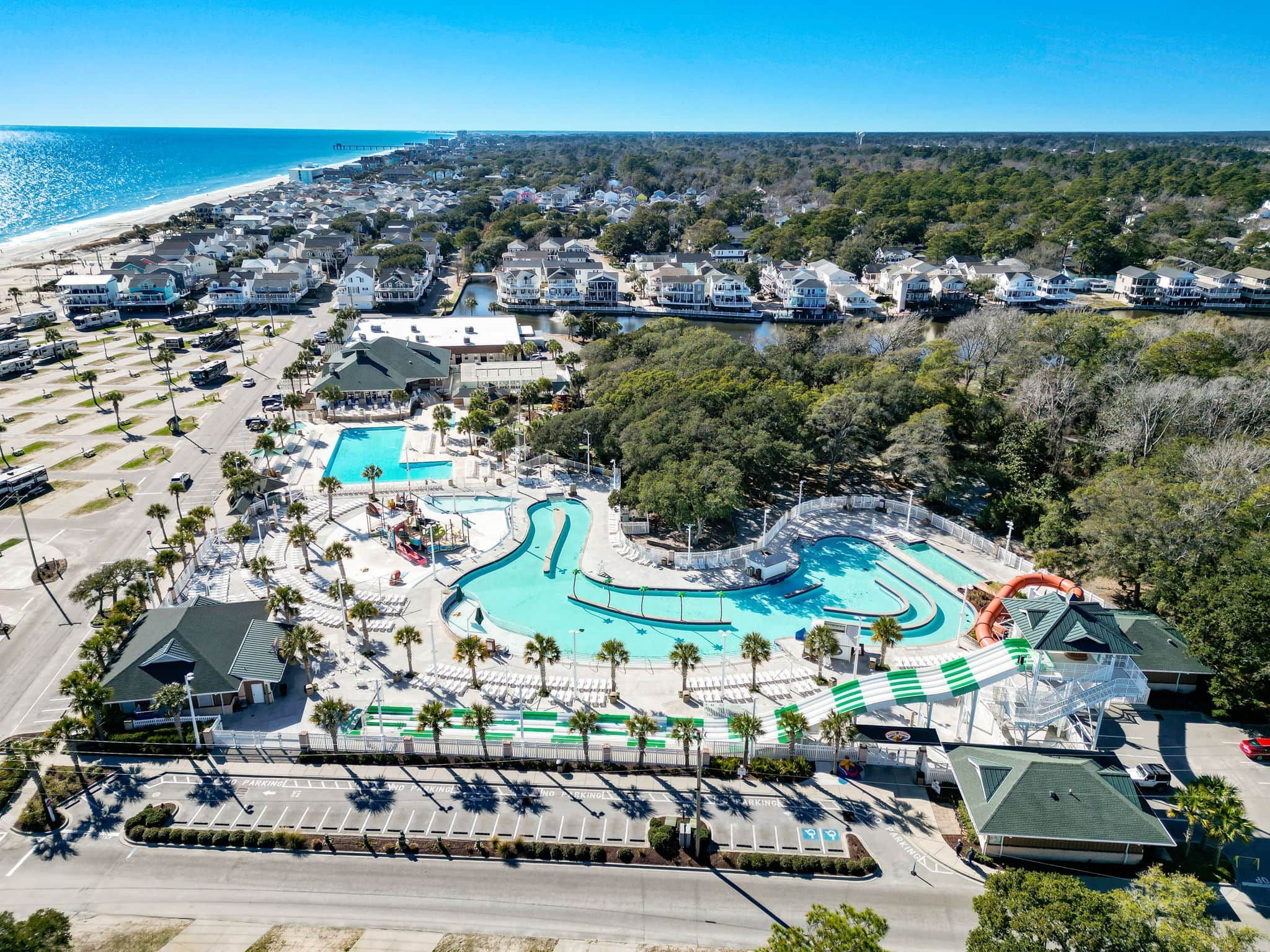 An aerial view of a resort with a large swimming pool, beachfront properties, and a parking area.