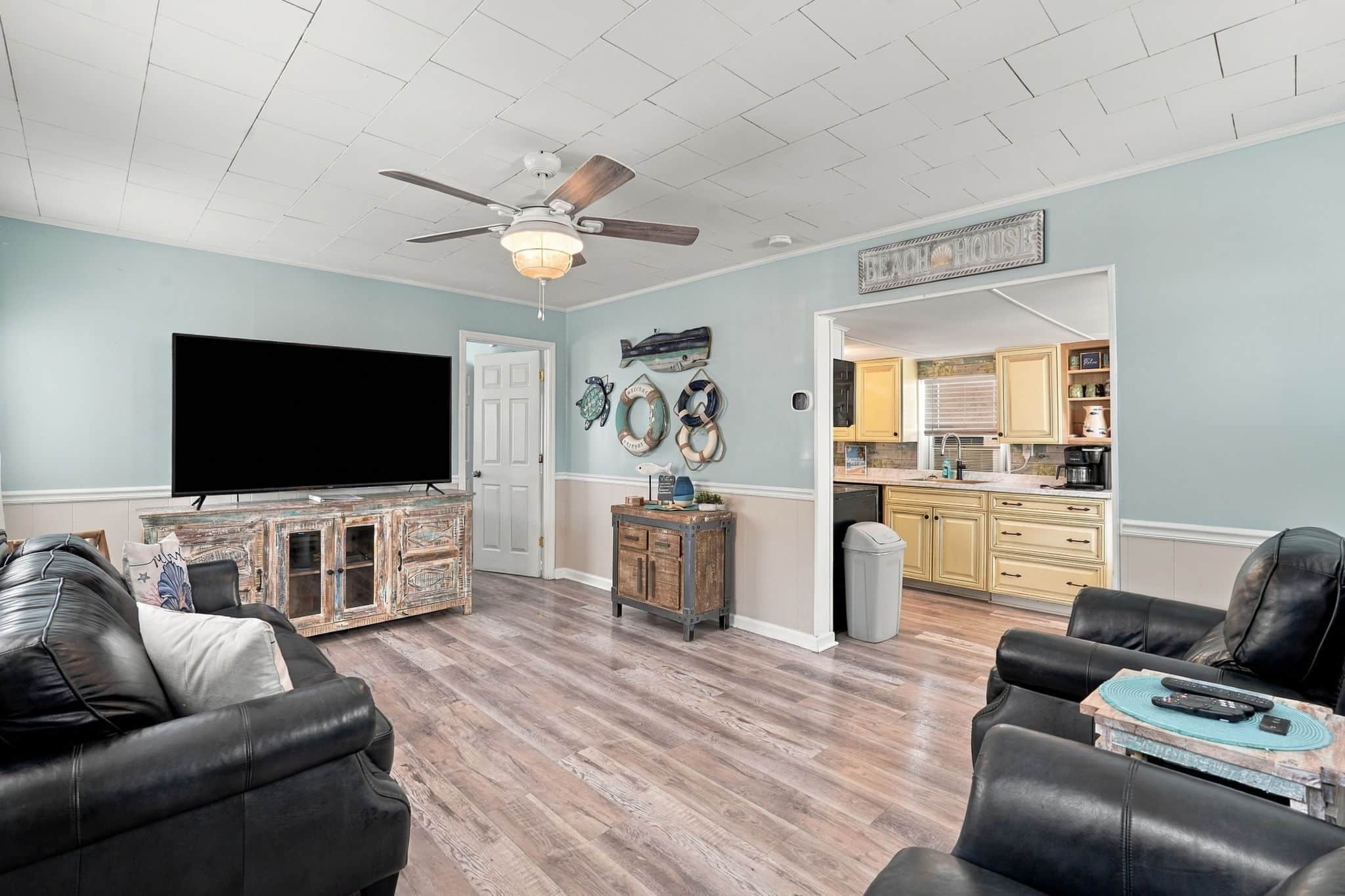 A living room with a flat-screen TV, leather sofas, a ceiling fan, and a kitchen visible through an open doorway.