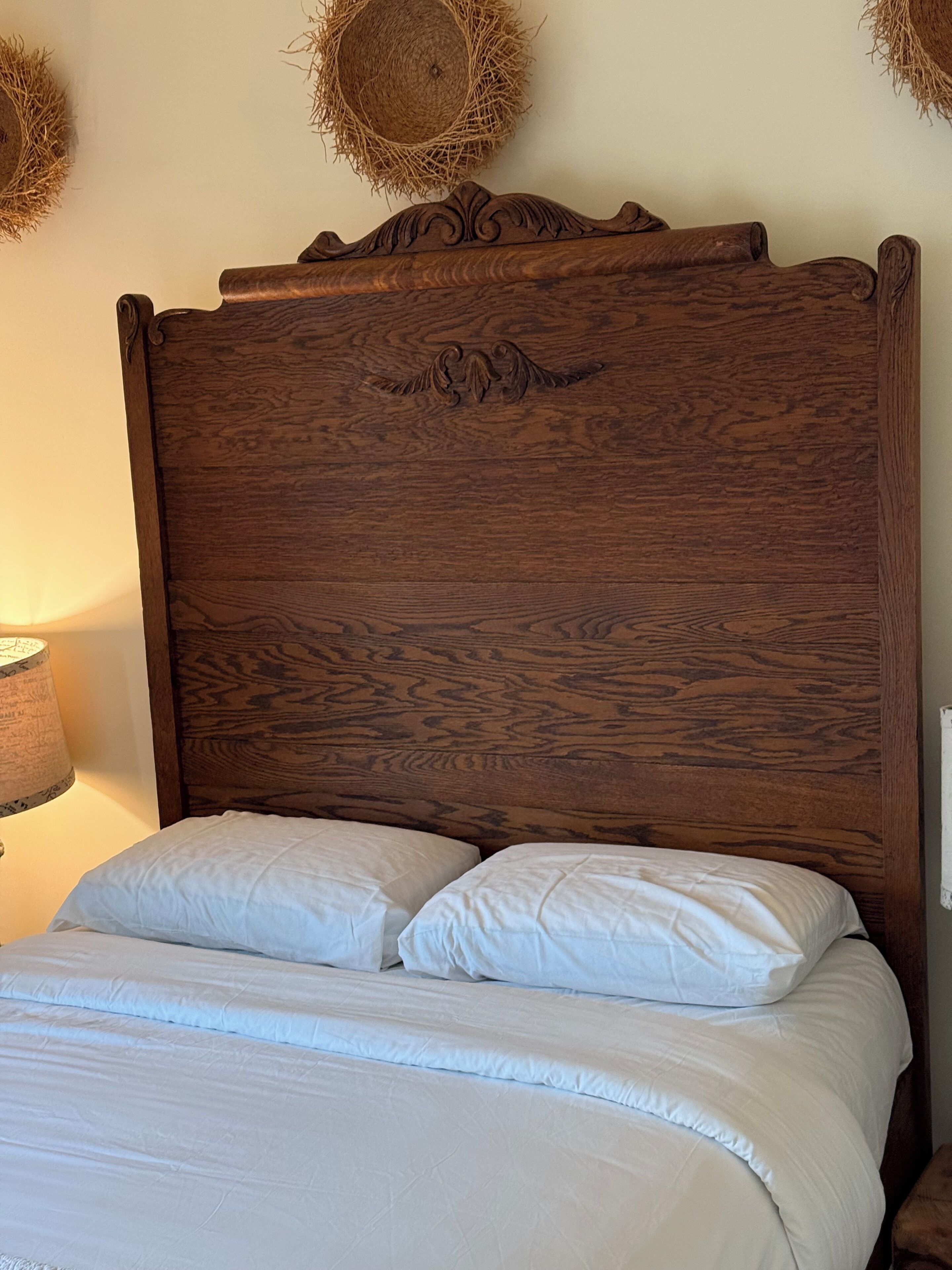 A wooden headboard with a carved design, a white bed with two pillows, and a lamp with a patterned shade on a bedside table.