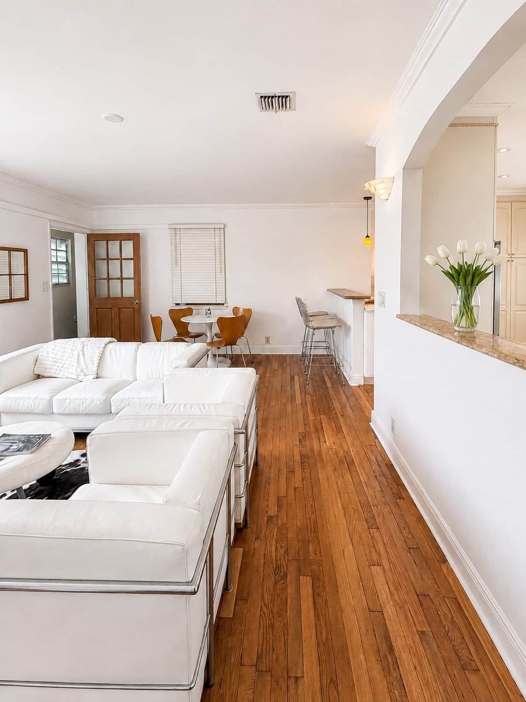 En moderne stue med en hjørnesofa, et spiseområde med bord og stole, og et køkkenområde med en bordplade og en vase med blomster.