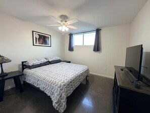 A bedroom with a bed, a ceiling fan, a window with curtains, a bedside table, and a TV on a stand.