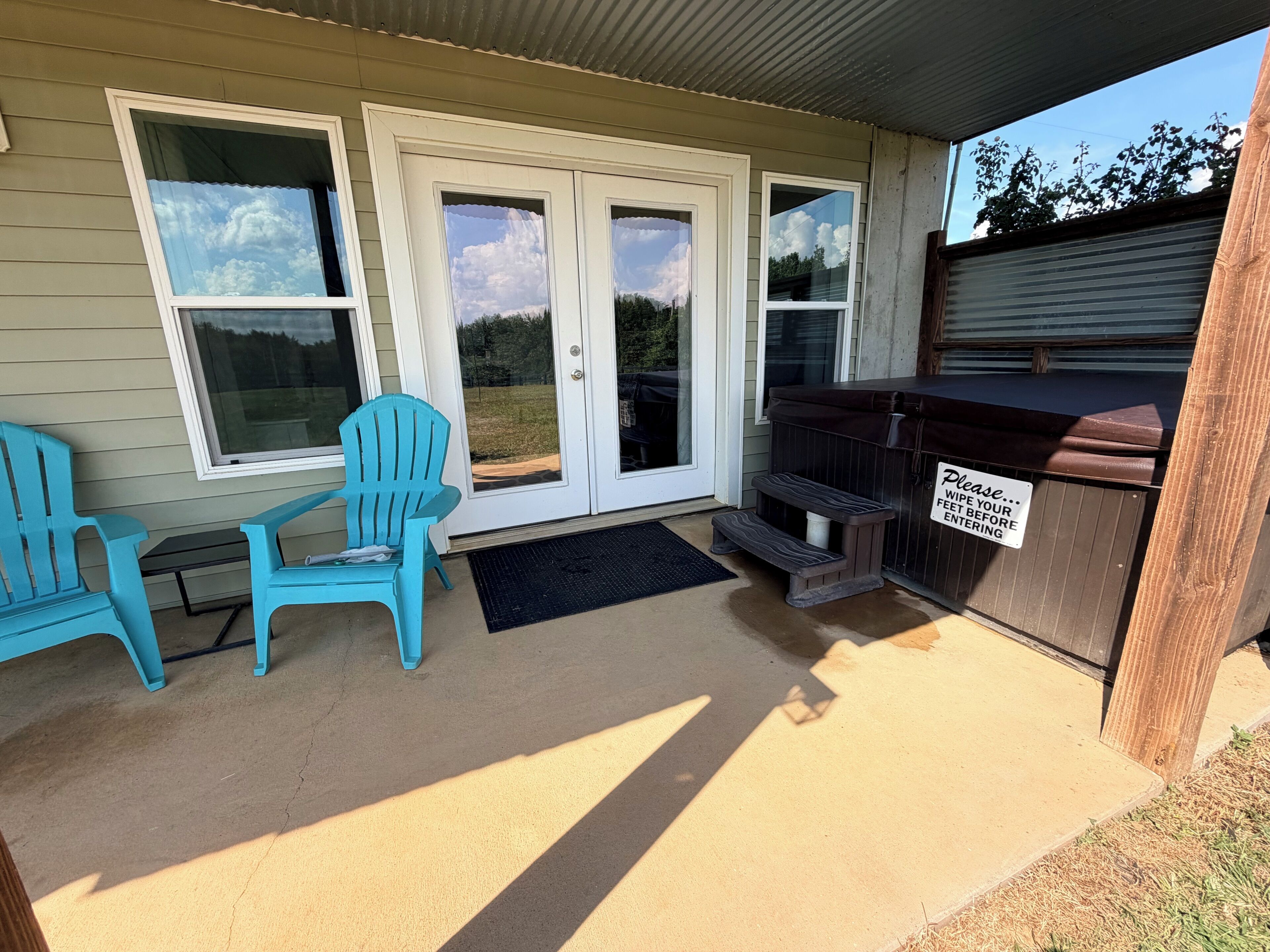 A covered outdoor area with two blue chairs, a barbecue grill, and a sign instructing to wipe feet before entering.